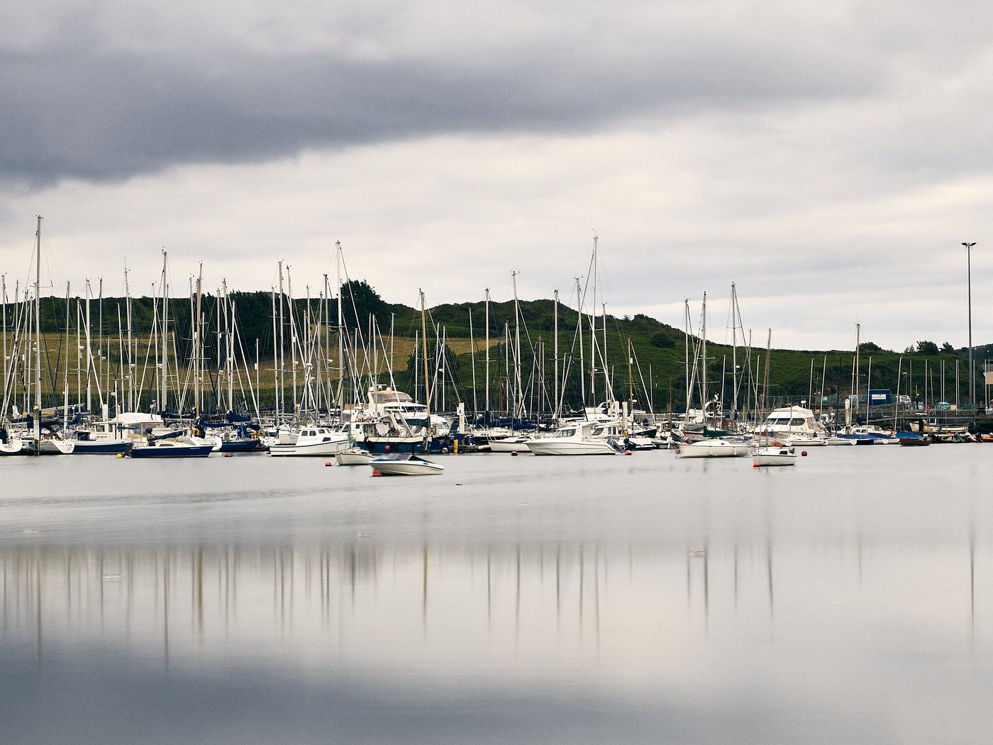 Boats moored on a calm estuary under a soft grey sky, green hills behind