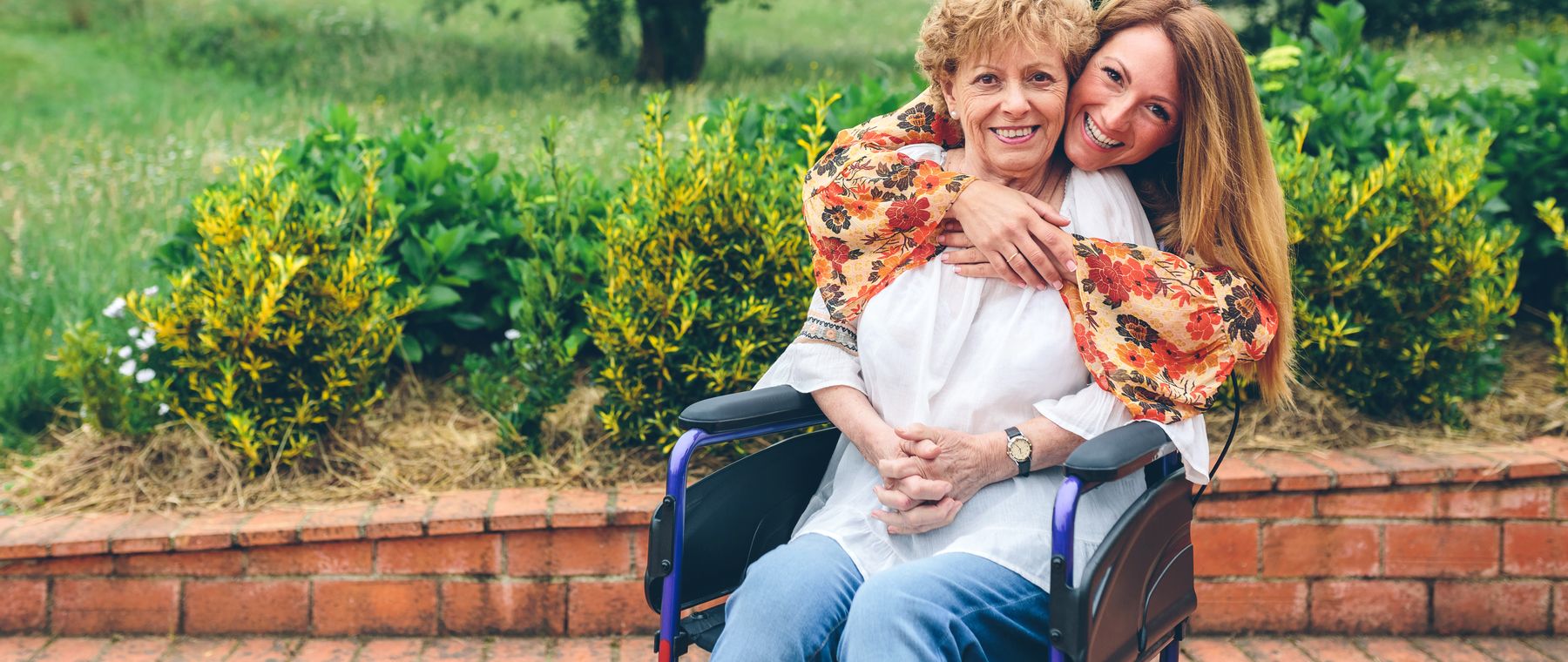 A daughter hugging her mother from behind in a sunny garden, her mother in a wheelchair and laughing