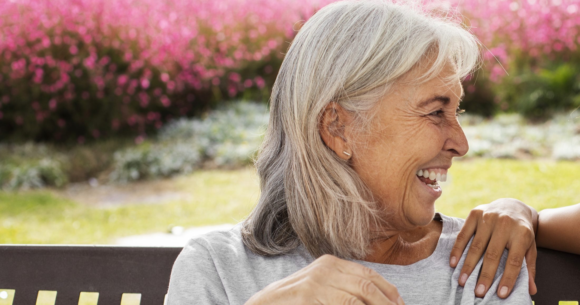 A resident laughing on the garden bench in the sunshine, a visitor's hand resting on her shoulder