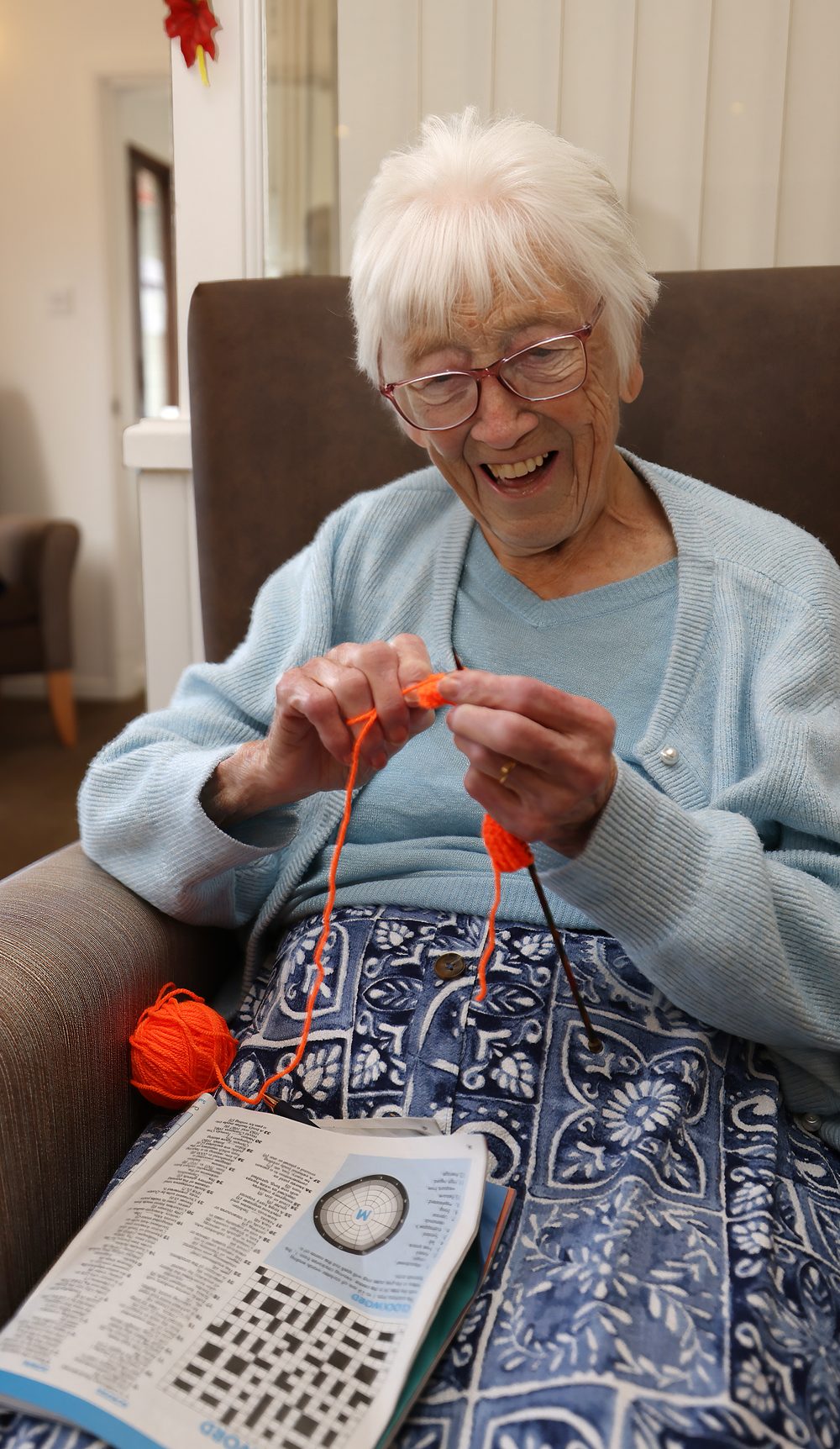 A resident smiling as she crochets in her armchair, a puzzle book on her lap
