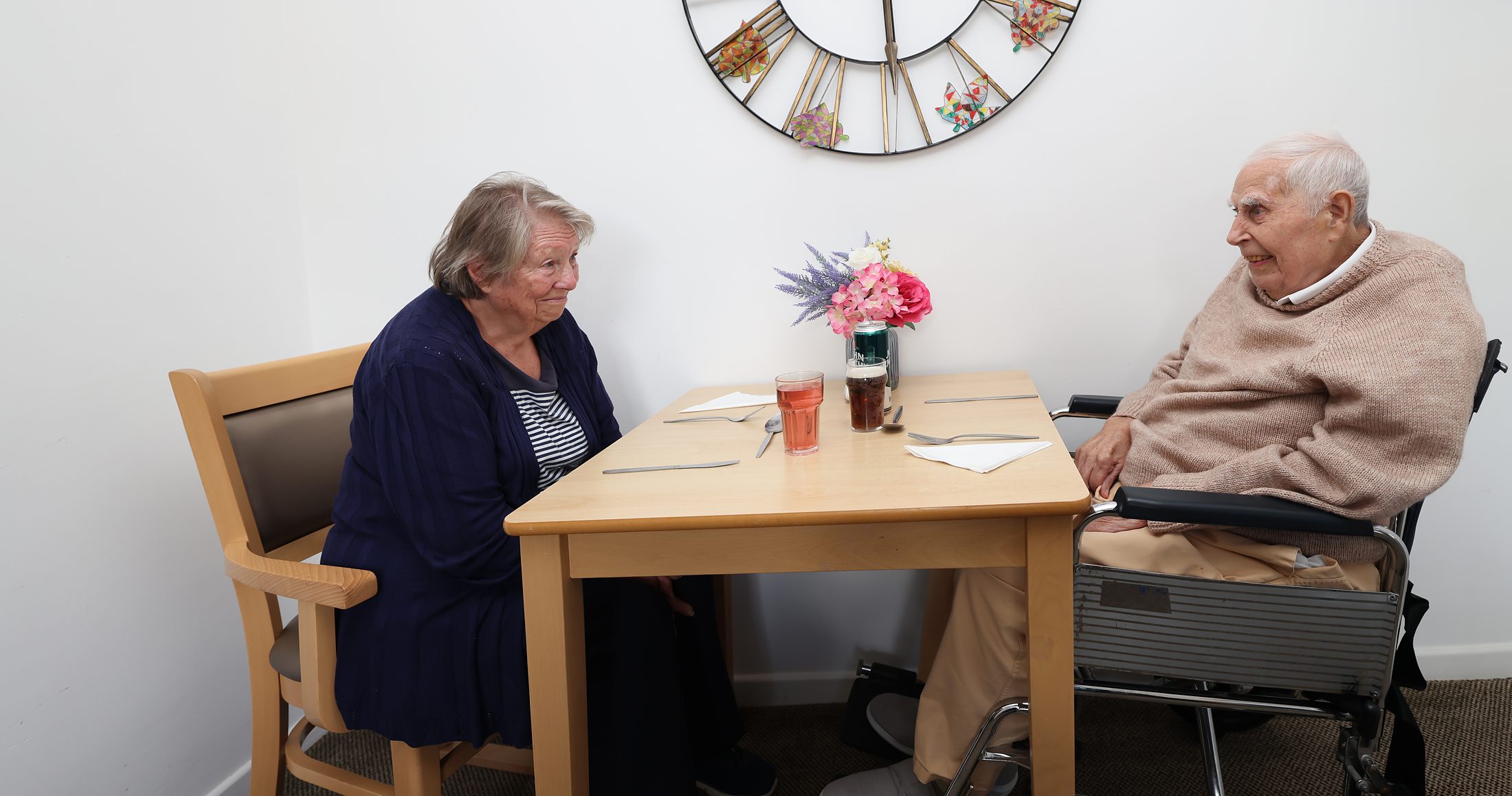 A resident and a family member chatting at a table, flowers between them